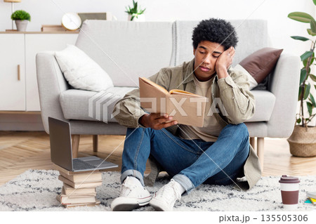 Distance education concept. Tired African American male student reading book near laptop at home. Black teenager sitting on floor with textbook, making his home assignment Distance education concept. Tired African American male student reading book near laptop at home. Black teenager sitting on floor with textbook, making his home assignment 135505306
