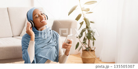 Full length portrait of pretty Muslim lady in hijab and headset listening to music on floor at home. African American woman in Arab scarf enjoying interesting audio book or nice melody 135505307