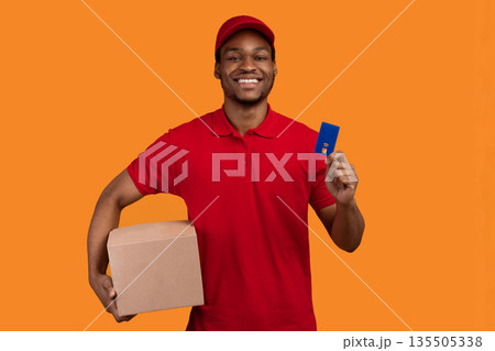 Easy Payment. Smiling african american male courier in red cap and uniform holding credit card and cardboard parcel box, carrying package, standing isolated over orange studio background Easy Payment. Smiling african american male courier in red cap and uniform holding credit card and cardboard parcel box, carrying package, standing isolated over orange studio background 135505338