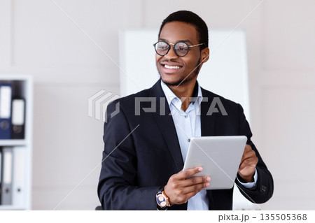 Modern gadgets and business. Joyful african american young businessman in suit using digital tablet in office, working online, getting ready for presentation, looking at copy space and smiling 135505368