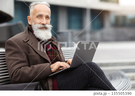 Wealthy grey-haired aged man with long beard in warm stylish outfit sitting on the bench by office building, using laptop, working online, chatting with clients or websurfing, empty space Wealthy grey-haired aged man with long beard in warm stylish outfit sitting on the bench by office building, using laptop, working online, chatting with clients or websurfing, empty space 135505390