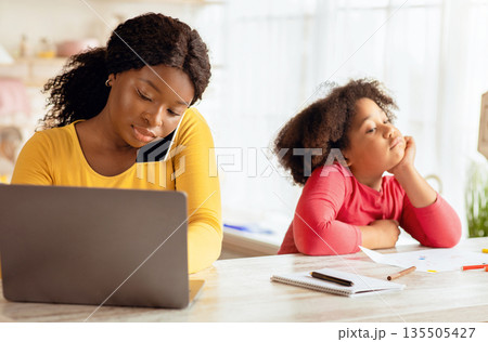 Portrait Of Busy Black Mom Working In Kitchen Next To Her Bored Little Daughter, Cute Female Child Feeling Lonely While Mother Using Laptop And Talking On Cellphone, Managing Remote Business Portrait Of Busy Black Mom Working In Kitchen Next To Her Bored Little Daughter, Cute Female Child Feeling Lonely While Mother Using Laptop And Talking On Cellphone, Managing Remote Business 135505427