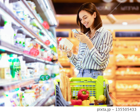 Smiling young woman taking dairy products from shelf in the supermarket, holding bottle and smartphone, scanning bar code on product through mobile phone, walking with trolley cart Smiling young woman taking dairy products from shelf in the supermarket, holding bottle and smartphone, scanning bar code on product through mobile phone, walking with trolley cart 135505433