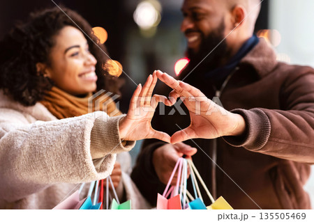 Romance And Bonding Concept. Portrait of happy smiling black african american man and woman lovers making heart shape with hands walking together in the evening after shopping, holding bags Romance And Bonding Concept. Portrait of happy smiling black african american man and woman lovers making heart shape with hands walking together in the evening after shopping, holding bags 135505469