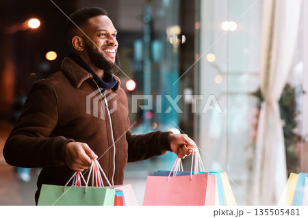 Portrait of smiling african american man looking at fashion clothes in shop window of the mall, walking in the evening near shopping center. Winter Gifts, Sales And Discount, Retail, Purchase 135505481