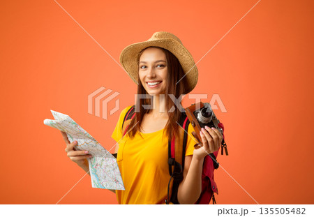 Happy young woman backpacker with camera holding city map over yellow studio background. Joyful lady traveller in summer hat looking at camera and smiling, going for sightseeing abroad, copy space Happy young woman backpacker with camera holding city map over yellow studio background. Joyful lady traveller in summer hat looking at camera and smiling, going for sightseeing abroad, copy space 135505482
