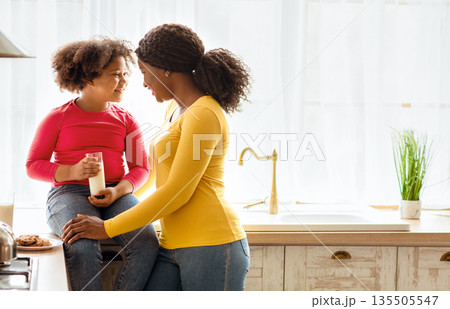 Happy black mom and daughter spending time together in kitchen, drinking milk and eating cookies. Cheerful little african american girl and her mother smiling and bonding at home, copy space 135505547