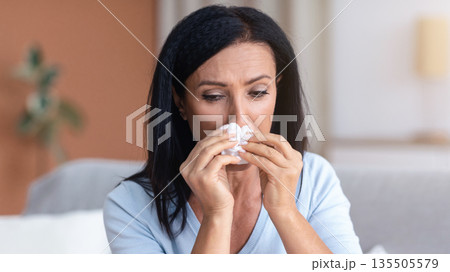 Portrait of mature brunette woman crying and sneezing, holding tissue paper napkin, sitting on the sofa at home. Sad middle-aged lady got cold or flu, feeling sorrow, suffering from runny nose 135505579