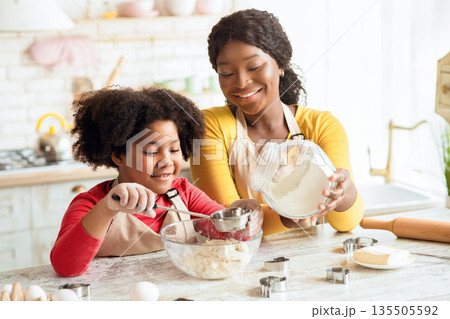 Happy african mother and her little daughter preparing dough for cookies together, adding flour and having fun in kitchen, doing homemade pastry, cute african girl and mom cooking at home, free space 135505592