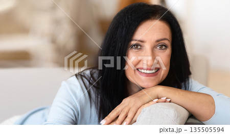 Closeup portrait of lovely middle-aged brunette woman smiling, sitting on sofa, leaning. Happy beautiful mature lady resting and relaxing at home looking at the camera, blurred background 135505594