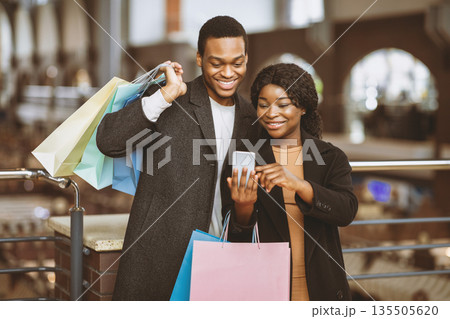 Super sale in stores in mall during Black Friday and autumn seasonal sale. Happy african american young male and female holds many color bags and makes selfie or watch stocks in smartphone, free space 135505620