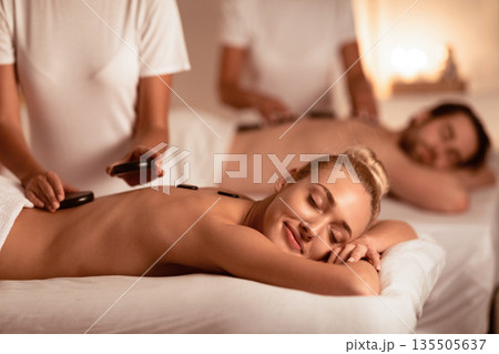 Exotic Relaxation. Spouses Receiving Hot Stones Massage Relaxing Lying On Beds At Luxury Spa Center, With Eyes Closed. Romantic Honeymoon Vacation. Low Light, Selective Focus 135505637