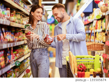 Millennial Family Couple Shopping Groceries In Supermarket Buying Food Walking With Trolley In Grocery Shop Indoor. Cheerful Customers Buy Supply Essentials Standing Near Shelves In Store Aisle 135505674