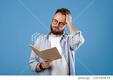 Young Caucasian student having difficulty understanding textbook, studying for exam on blue studio background. Puzzled millennial guy scratching head while reading complicated book 135505679