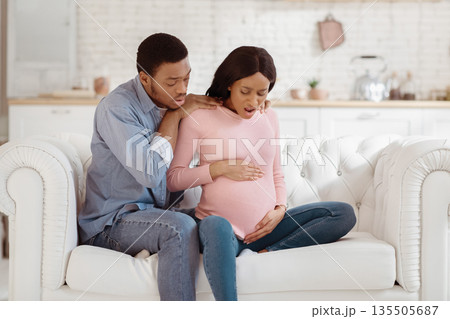 Pregnant black woman feeling acute pain, giving birth to child, having contractions while worried husband calling doctor on phone. African American lady in labor with her partner at home Pregnant black woman feeling acute pain, giving birth to child, having contractions while worried husband calling doctor on phone. African American lady in labor with her partner at home 135505687