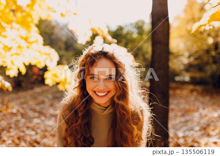 Smiling young woman enjoys the autumn weather in the forest with the yellow leaves at sunset Smiling young woman enjoys the autumn weather in the forest with the yellow leaves at sunset 135506119