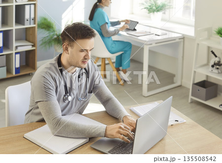 Portrait of a young male doctor in uniform sitting at the desk and working in office 135508542