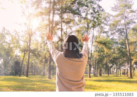 Rear view of a woman raising her hands in a sunny forest park at sunrise. Concept of gratitude, worship, prayer, freedom, mindfulness, healing and mental wellbeing in nature. Rear view of a woman raising her hands in a sunny forest park at sunrise. Concept of gratitude, worship, prayer, freedom, mindfulness, healing and mental wellbeing in nature. 135511083