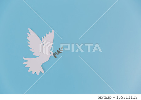 White paper-cut dove holding an olive branch on a blue background with ample copy space. Minimal peaceful symbol for hope, freedom, faith, unity and humanitarian concepts. 135511115