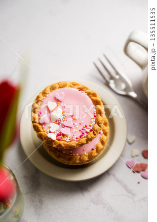 Valentine's day. Pink mini tarts with heart shaped sprinkles, cup of coffee and bouquet of spring red tulips Valentine's day. Pink mini tarts with heart shaped sprinkles, cup of coffee and bouquet of spring red tulips 135511535