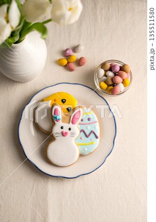 Easter festive background. Colourful easter cookies bunny, egg and chick in a plate with eggs candies and bouquet of white spring tulips 135511580
