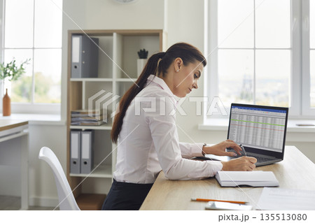 Focused woman managing finances at home office with laptop and notebook, budgeting Focused woman managing finances at home office with laptop and notebook, budgeting 135513080