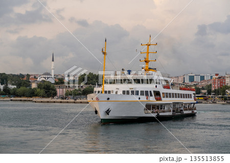A ferry navigates the Bosphorus Strait, gliding past the vibrant city skyline, historic structures, and lush greenery under a moody sky. 135513855