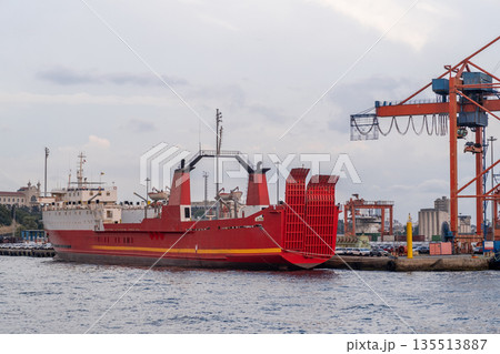 A large red cargo ship is moored at a dock along the Bosphorus strait as cranes overlook the bustling port at dusk, creating a busy maritime atmosphere. 135513887