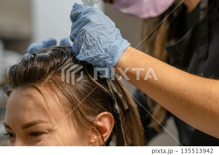 A woman sits in a salon chair as a stylist applies hair color. The atmosphere is relaxed, focusing on the hair transformation. 135513942
