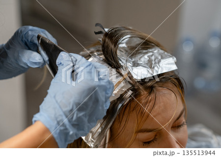 Stylist applies hair dye using foils while client relaxes in a well lit salon setting in the afternoon. 135513944