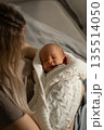 A mother gently holds her serene baby swaddled in a soft blanket, creating a peaceful atmosphere in a dimly lit room during evening hours. 135514050