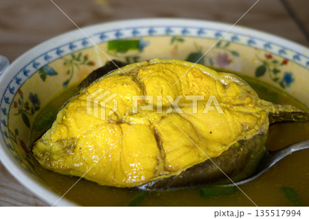 Close-up of ikan sembilang asem asem or Indonesian sour soup of Eel-tailed Catfish dish in a floral bowl with a spoon, on a wooden table with warm tones. 135517994