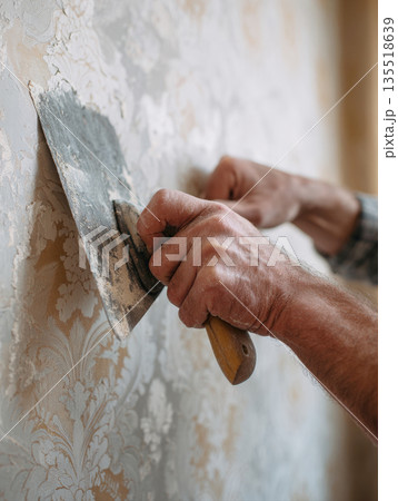 Skilled Worker's Hands Using a Wide Metal Spatula to Scrape and Remove Old, Patterned Wallpaper from a Wall During Home Renovation, Repair, DIY Decorating Projects, Illustrating the Preparation Stage 135518639