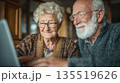 a cheerful senior woman and man with a white beard, sitting closely together as they browse or video chat on a computer, highlighting their connection and technological literacy. 135519626