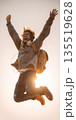 A low-angle action photograph capturing a cheerful, energetic traveler or student mid-jump, wearing headphones around his neck, expressing pure happiness, freedom, and exuberance against a bright sky. 135519628