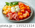 Colorful salad with pieces melon, prosciutto meats and mozzarella served on gray plate with fresh basil on white concrete background 135521314