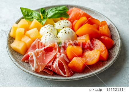 Colorful salad with pieces melon, prosciutto meats and mozzarella served on gray plate with fresh basil on white concrete background 135521314