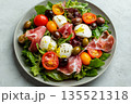 Top view of fresh salad with prosciutto meat, greens, cherry tomatoes, olives, mozzarella cheese and olive oil served on gray plate on concrete background, delicious and healthy dish 135521318