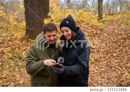Man and woman standing close together looking at smartphone screen in autumn park 135521886