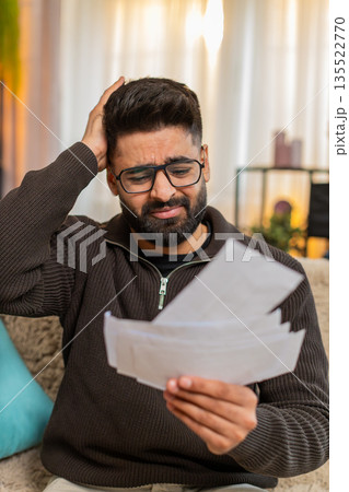 Indian man looking through receipts with worry on sofa at home, financial stress and anxiety visible 135522770