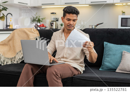 Indian man updates bill numbers in laptop spreadsheet checks docs and confirms monthly budget total Indian man updates bill numbers in laptop spreadsheet checks docs and confirms monthly budget total 135522781