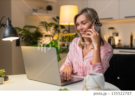 Adult woman works on laptop talks on smartphone multitask productive remote clear voice focus call Adult woman works on laptop talks on smartphone multitask productive remote clear voice focus call 135522787