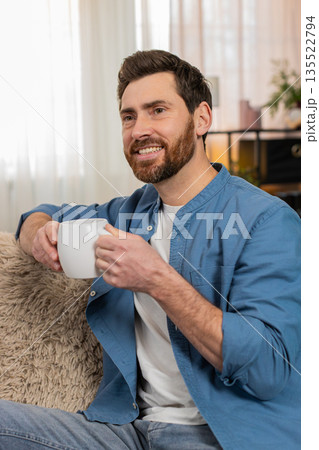 Young man sitting on sofa holding a cup drinking tea, gazing into distance enjoying the moment 135522794