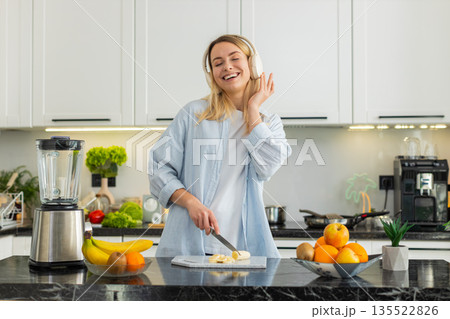 Young woman slicing banana for smoothie blender while dancing with headphones for healthy fit diet 135522826