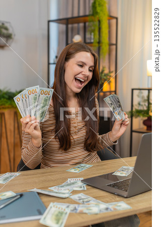 Young woman wins online casino jackpot on laptop celebrates and holds dollar bills as cash on table Young woman wins online casino jackpot on laptop celebrates and holds dollar bills as cash on table 135522829