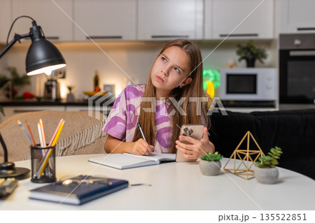 Teenager girl online math lesson on smartphone serious focus writes notes in notebook for schoolwork Teenager girl online math lesson on smartphone serious focus writes notes in notebook for schoolwork 135522851