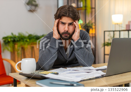 Indian man holds head over unpaid bills on table with calculator and laptop worried about money debt 135522906