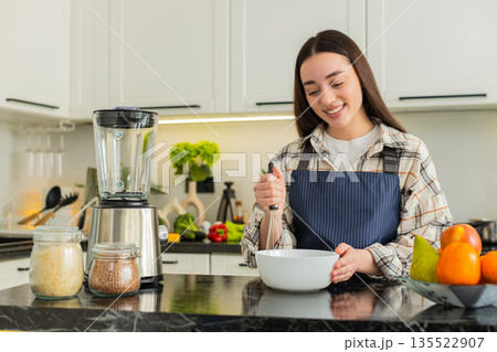 Young woman whisking ingredients in kitchen preparing homemade meal recipe for healthy fit nutrition 135522907