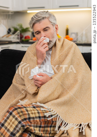 Middle-aged ill man under plaid sneezes uses tissue sniffles sick flu virus home quarantine rest Middle-aged ill man under plaid sneezes uses tissue sniffles sick flu virus home quarantine rest 135522943