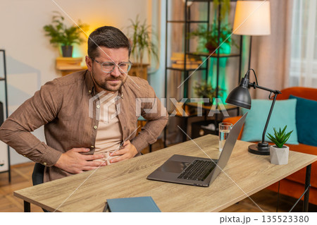 Disappointed freelancer man feeling stomach ache overeating during work process on laptop at home Disappointed freelancer man feeling stomach ache overeating during work process on laptop at home 135523380
